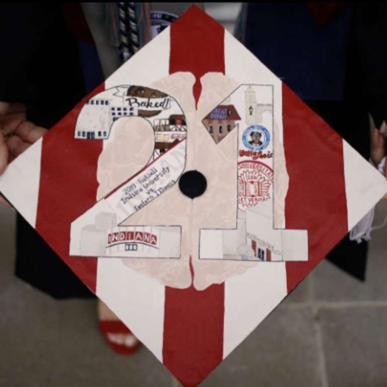 A decorated mortarboard with red and white stripes and illustrations of the graduate's favorite things in the numbers "21."