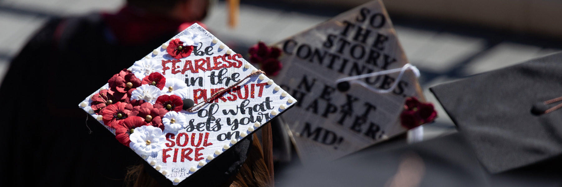 Graduate seen from behind with mortarboard decorated with red and white flowers and decorative writing that says: Be fearless in the pursuit of what sets your soul on fire.