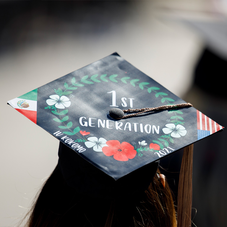 A young woman wears a decorated mortarboard with the colors of the US and Mexican flags and text saying "first generation"