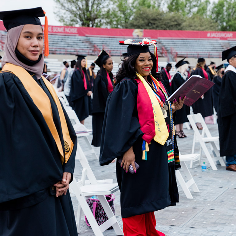 Two young women graduating from Purdue schools and IU schools of IUPUI smile for the cameras at IUPUI's outdoor spring commencement.